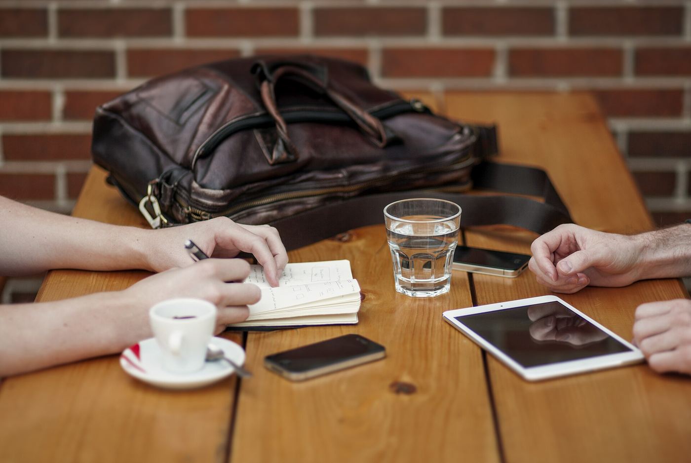 Two people at a wooden cafe table with an iPhone, a second phone, an iPad and a paper notebook between them