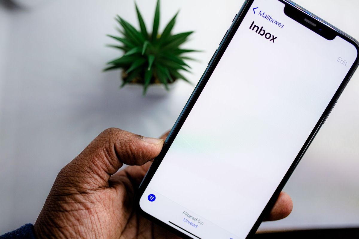 A hand holding an iPhone showing an empty email inbox in front of a potted plant