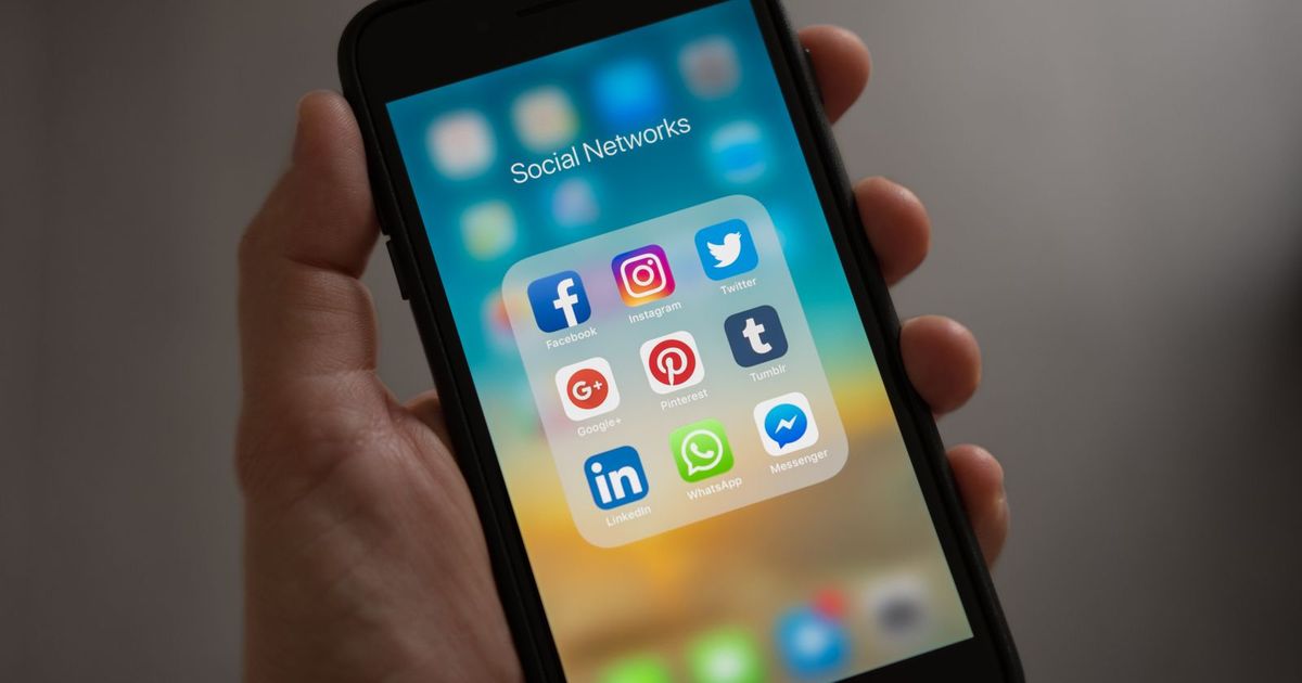 A hand holding an iPhone showing a home screen folder named Social Networks, with Facebook, Instagram, Twitter, Pinterest, LinkedIn and WhatsApp icons inside