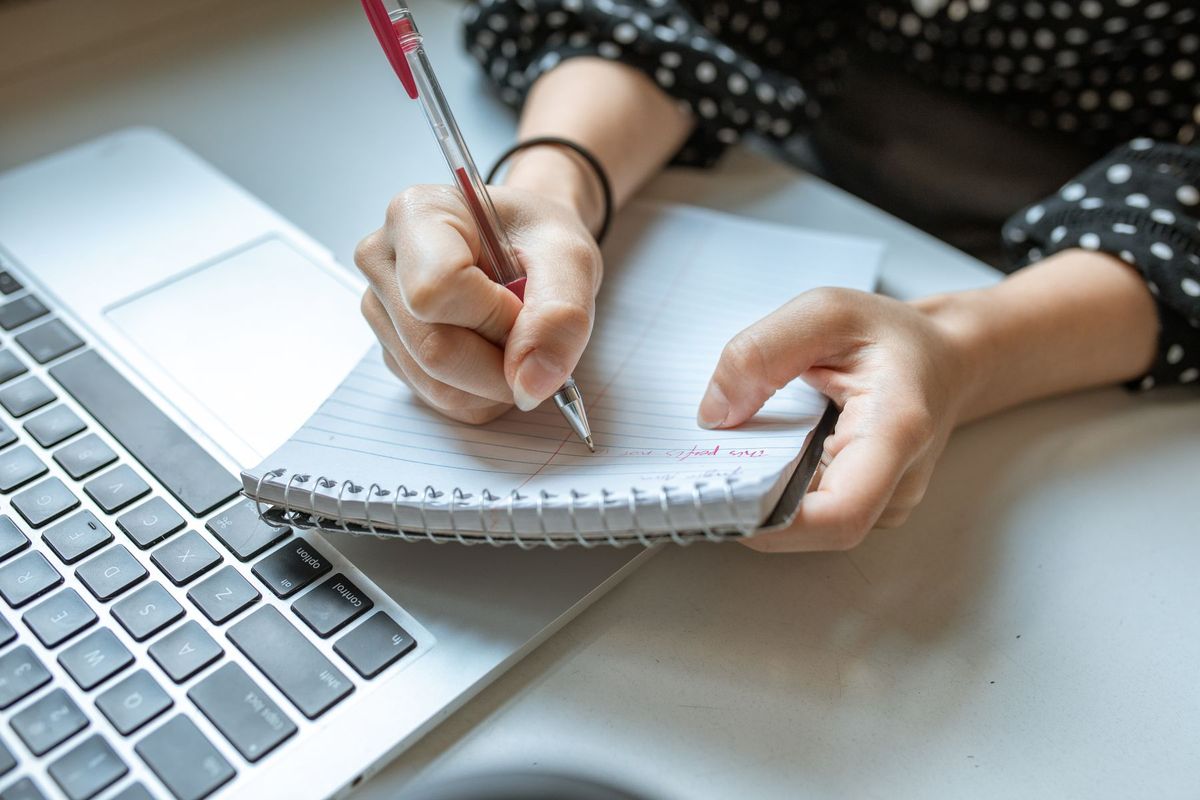 A person writing in a spiral notebook beside an open laptop