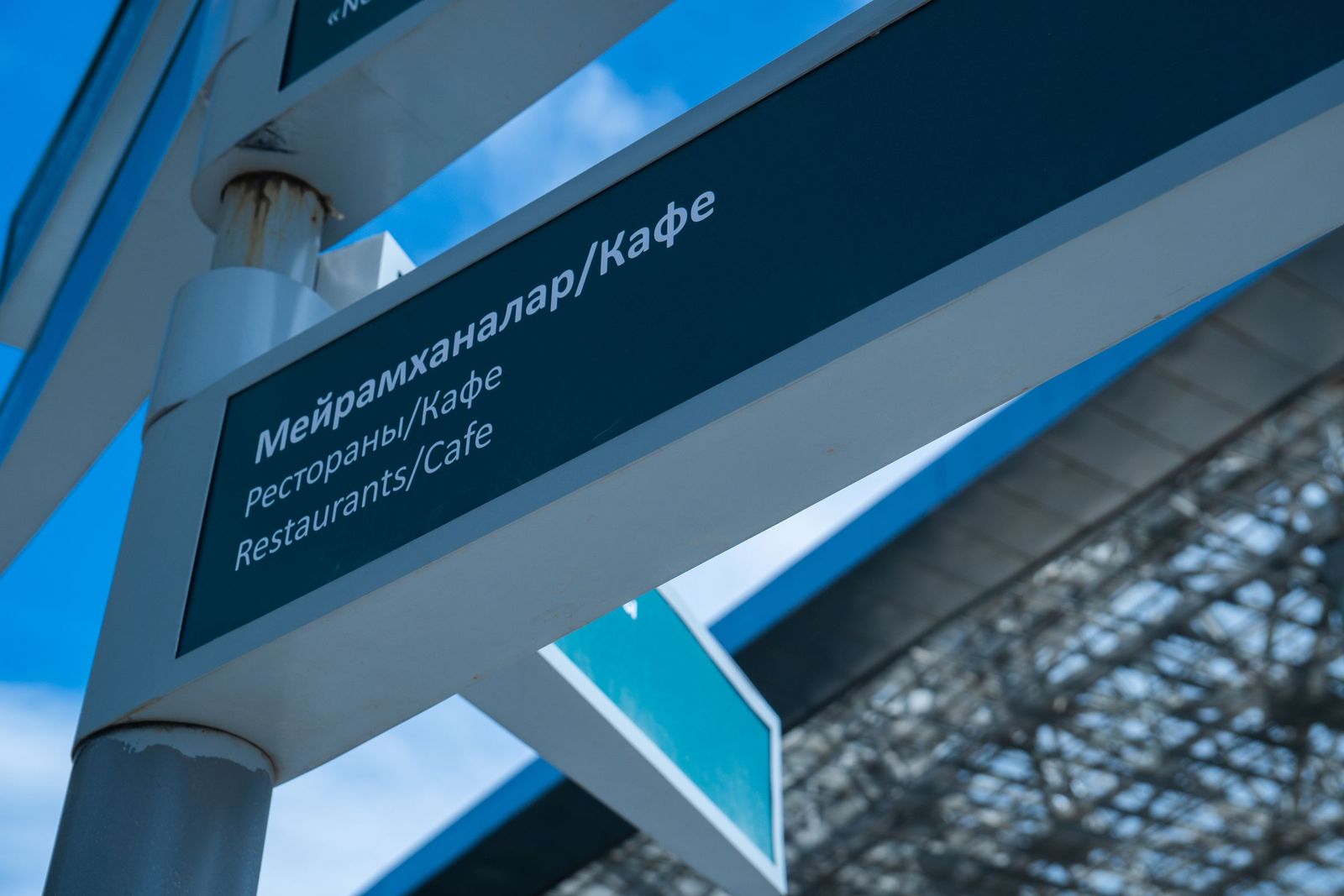 A close-up of a directional sign in Kazakh, Russian and English pointing to restaurants and cafes, against a blue sky