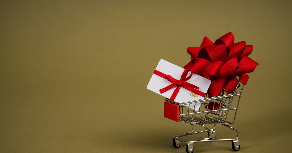 A white gift card tied with a large red ribbon bow sitting in a miniature shopping cart on an olive green background