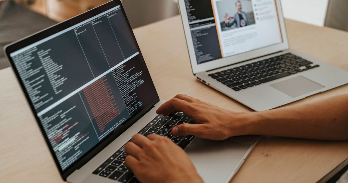 Hands typing on a MacBook showing a terminal full of code, with a second laptop open in the background on a wooden desk