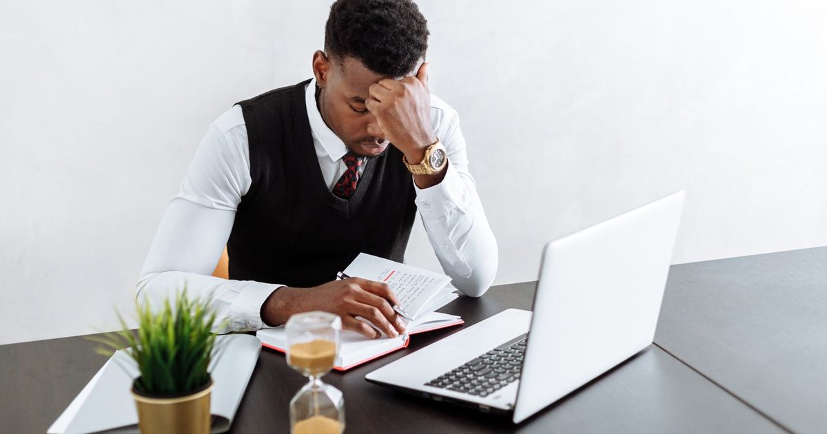A man at an office desk with his head in his hand, writing in a notebook next to an open laptop and a small hourglass
