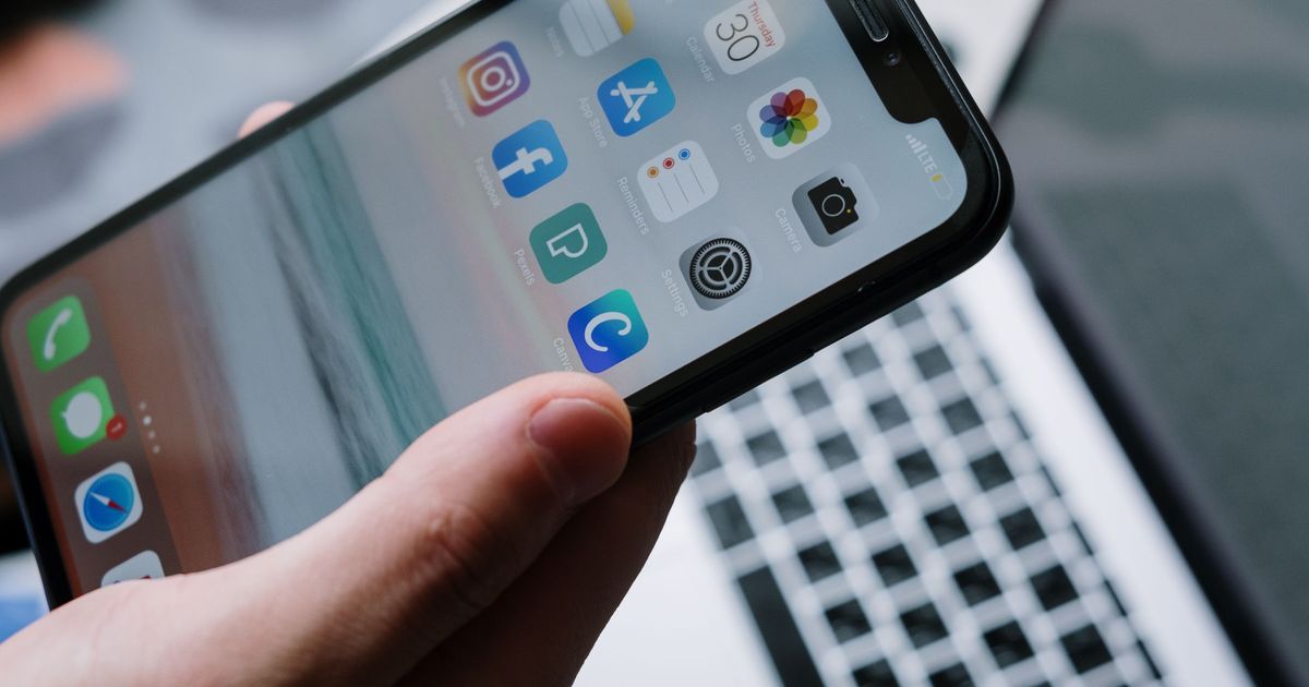 A hand holding an iPhone showing a home screen of app icons, including the App Store, in front of an open laptop keyboard