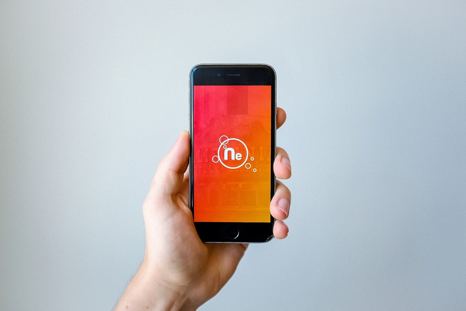 A hand holding up an iPhone whose whole screen shows an app's full-bleed orange launch artwork with its logo, against a plain grey wall