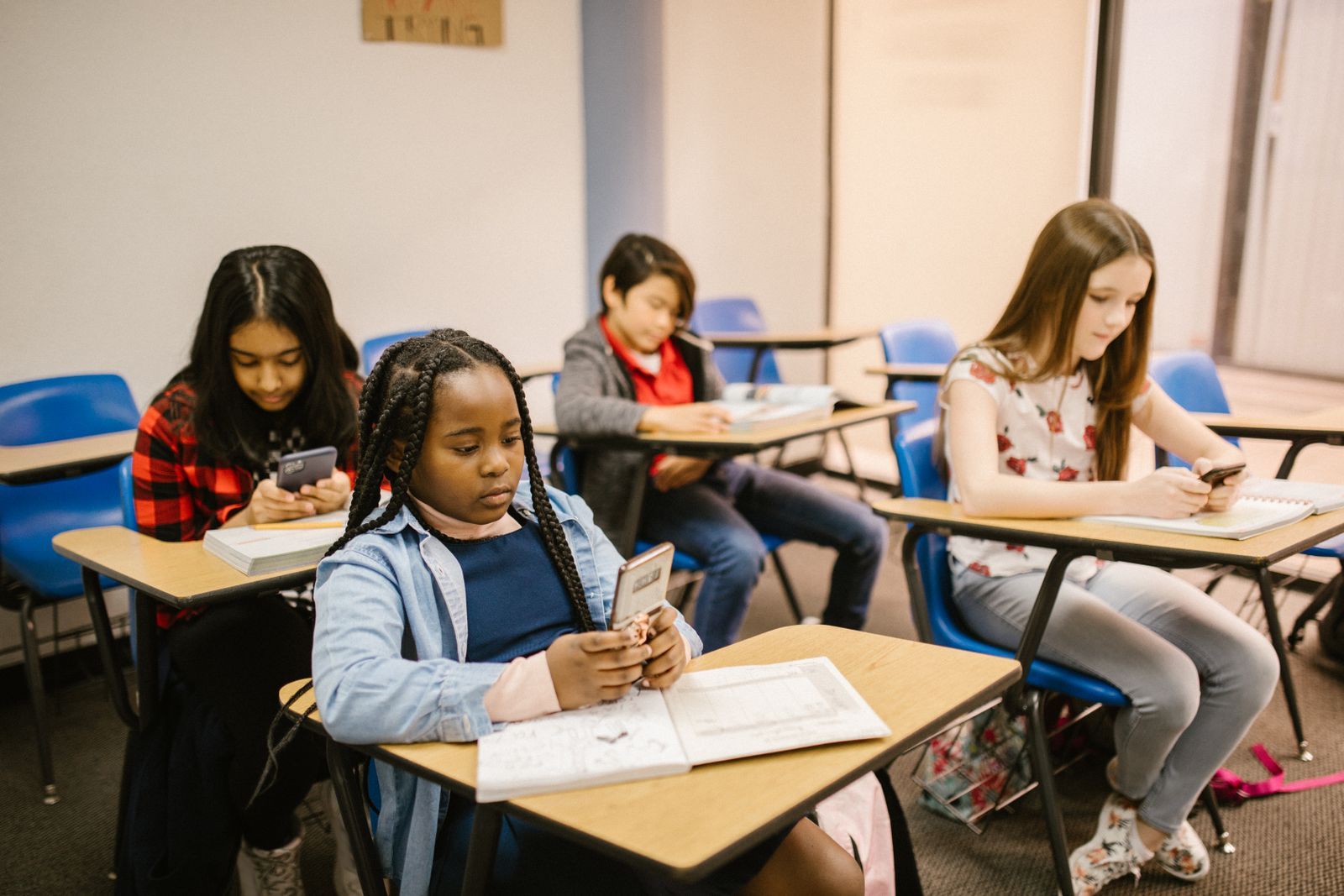Four school students at classroom desks, each looking down at a smartphone