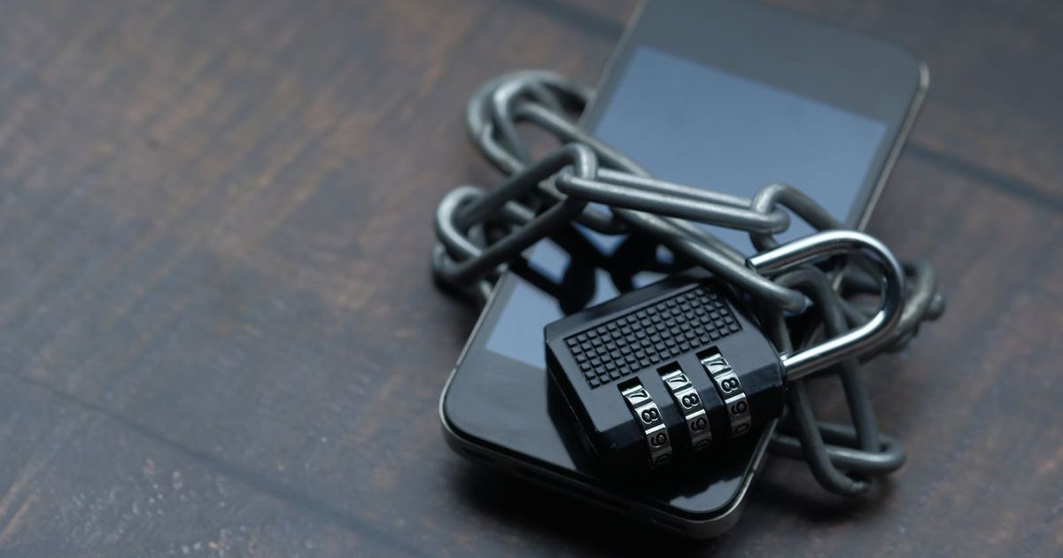 A black smartphone lying face up on a wooden table, wrapped in a steel chain and locked with a combination padlock