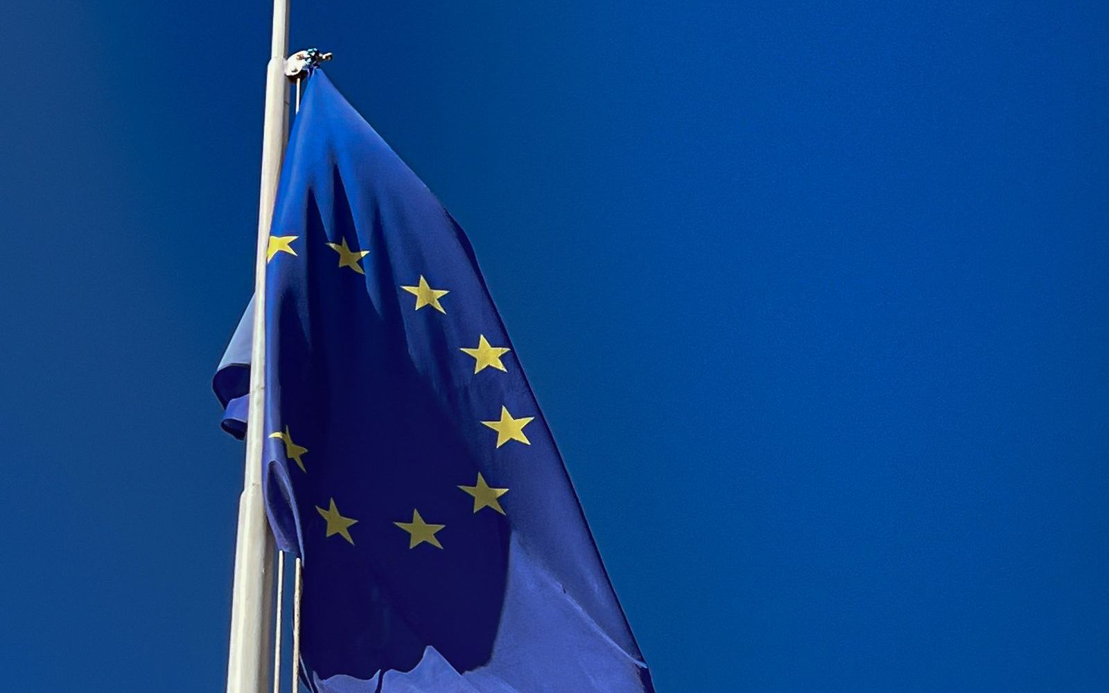 The blue European Union flag with its ring of yellow stars, hanging from a white flagpole against a clear blue sky