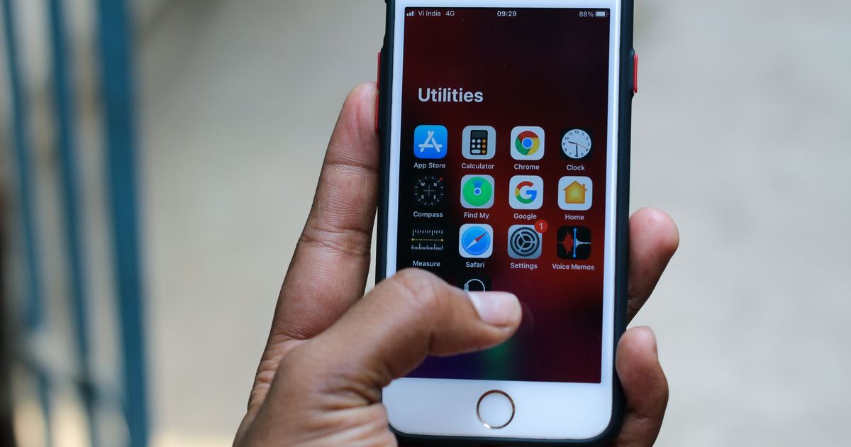 A hand holding an iPhone open on a Utilities folder, with the App Store icon in the top left corner of the folder