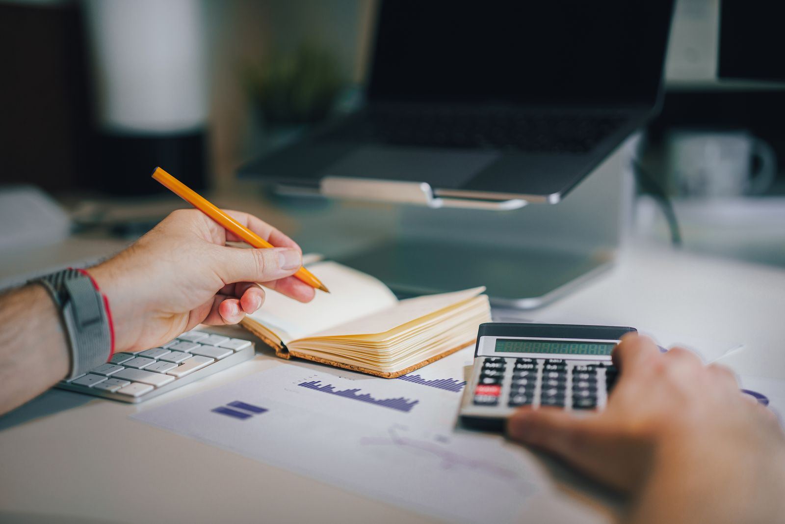 Hands writing in a notebook and using a calculator on top of printed charts, with a laptop behind them