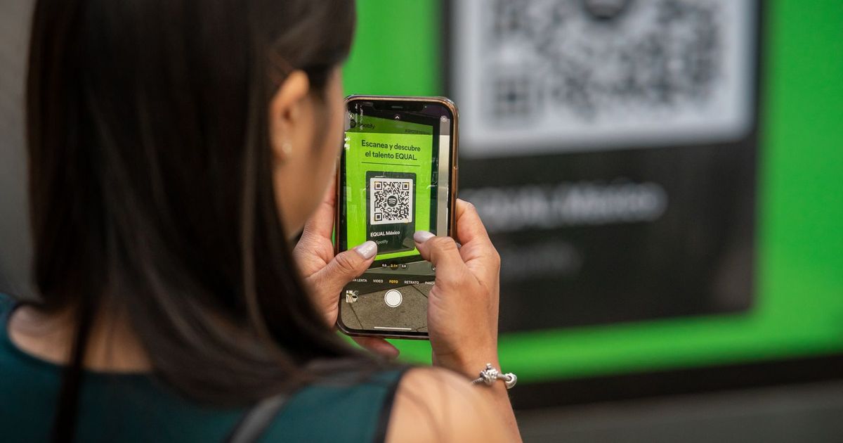A woman photographing a QR code on a green poster with her smartphone, the code visible on her phone screen
