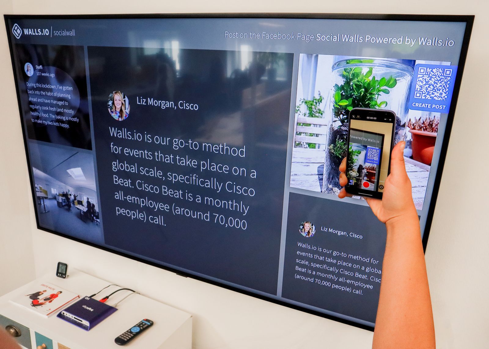 A hand holding a phone up to a large wall screen, scanning a QR code shown in the corner of the display