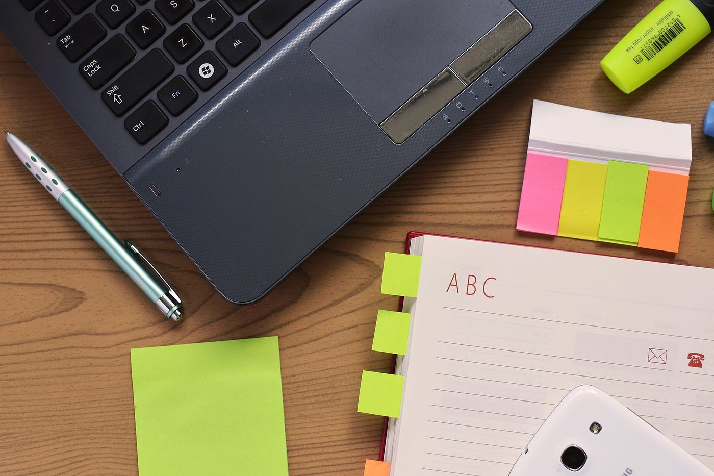 A laptop on a wooden desk with a pen, a highlighter, coloured page markers, sticky notes and an open notebook