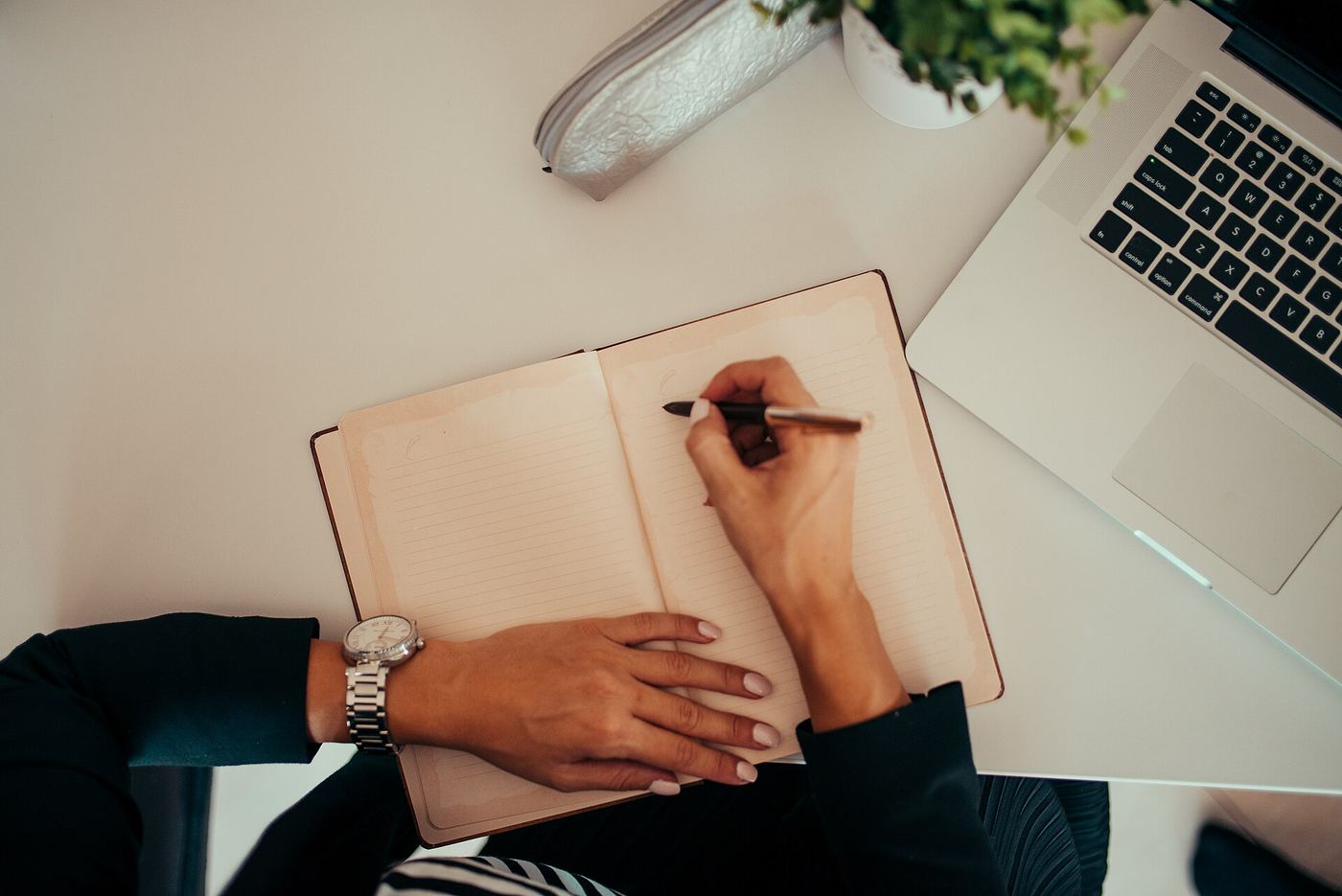 A person writing in an open notebook at a white desk, with a laptop and a small plant beside them