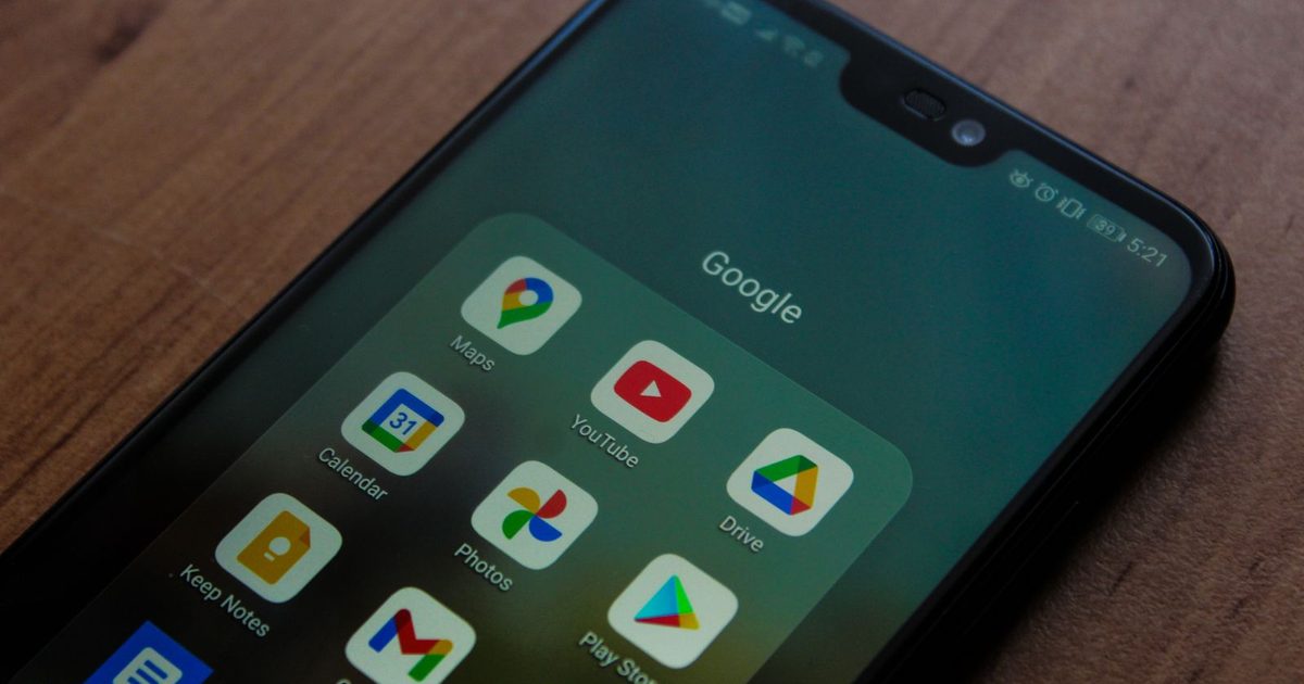 An Android phone on a wooden table with a Google folder open, showing Maps, YouTube, Calendar, Drive, Photos, Gmail and Play Store icons