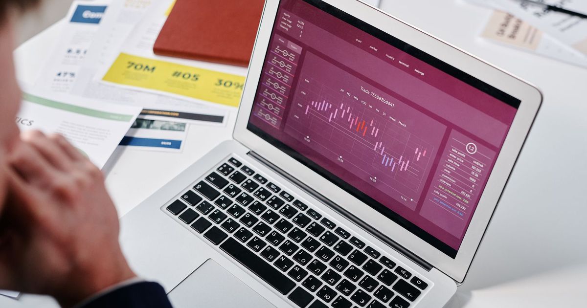 A laptop on a white desk showing a dark red analytics dashboard with a line chart, printed marketing reports with percentages spread around it