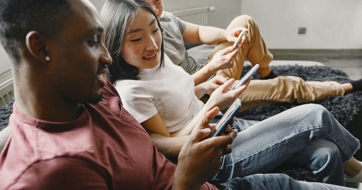 Three friends sitting together on a sofa, each looking at and tapping on their own smartphone