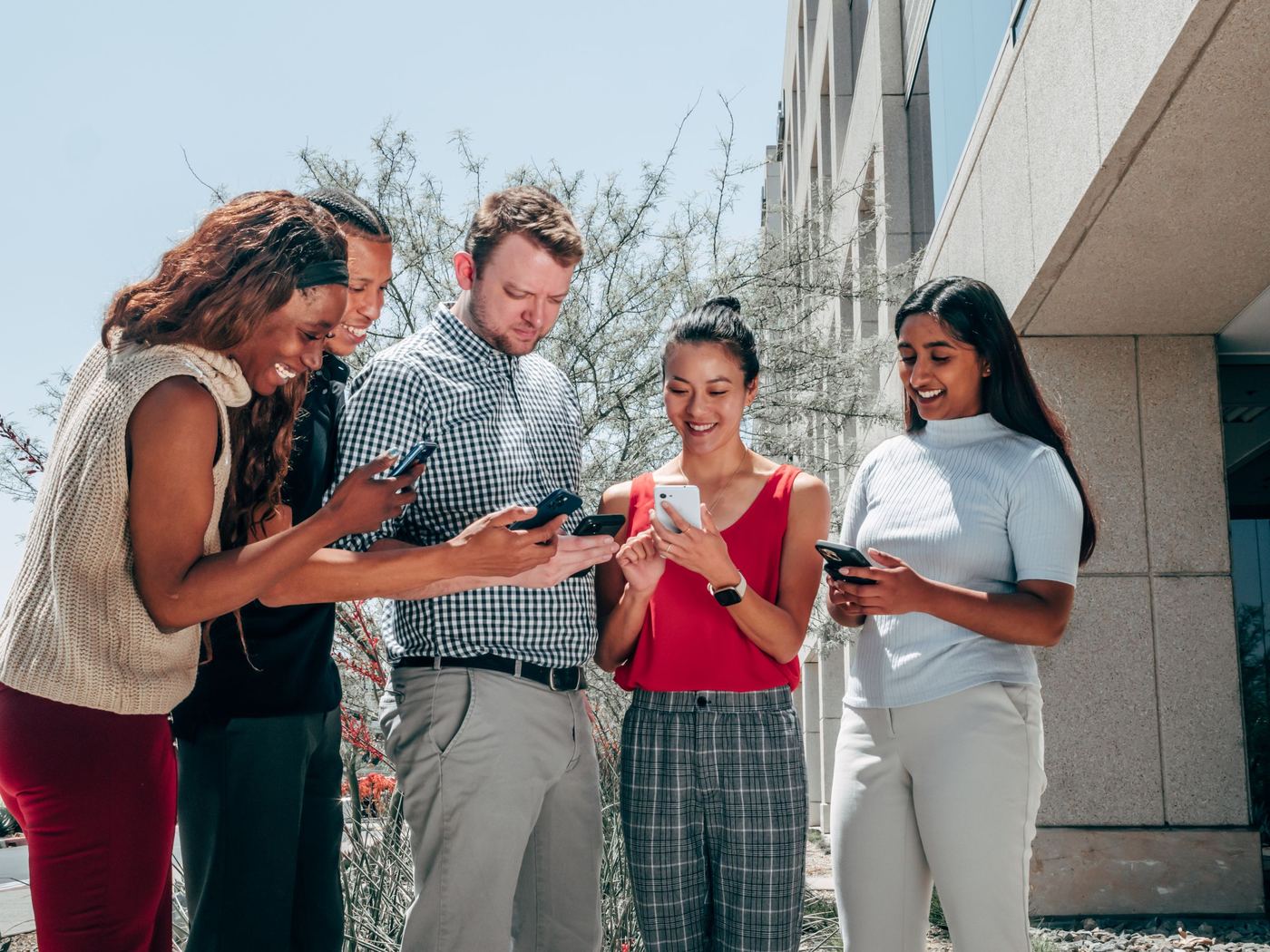 Five friends standing outdoors, each holding and using a smartphone