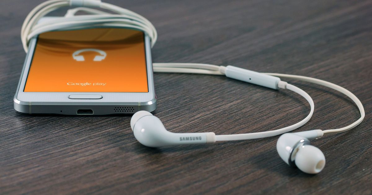 A white Samsung phone on a dark wooden table with earphones wrapped around it, its orange screen showing a headphones icon and the Google Play wordmark