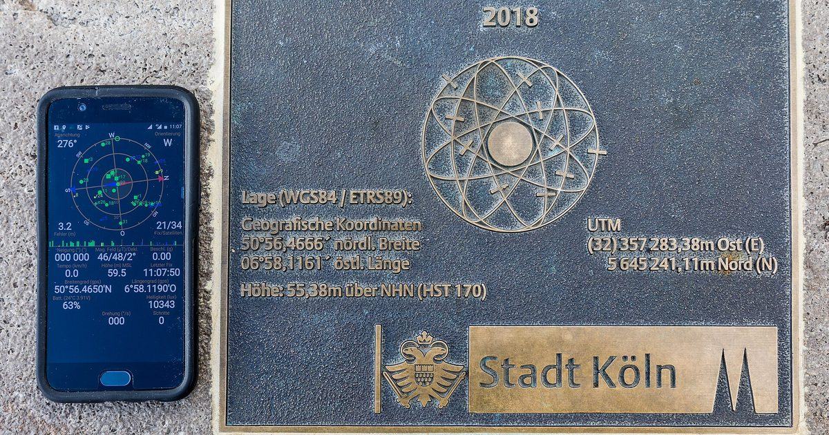 A smartphone showing a GPS satellite status app with coordinates, lying on paving stones beside a bronze GPS reference point plaque from the city of Cologne