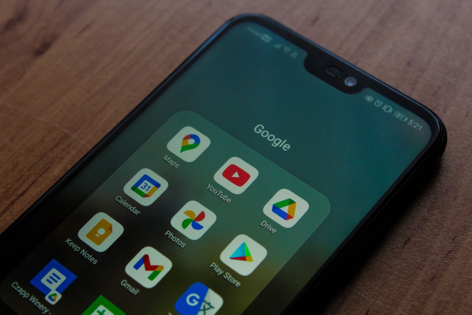 An Android phone on a wooden desk showing a Google folder with Maps, YouTube, Drive, Photos, Gmail and the Play Store icons