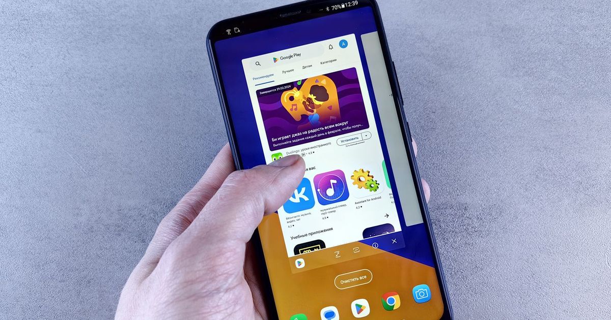 A hand holding an Android phone against a grey wall, its screen showing the Google Play Store app with a featured game banner and rows of app icons