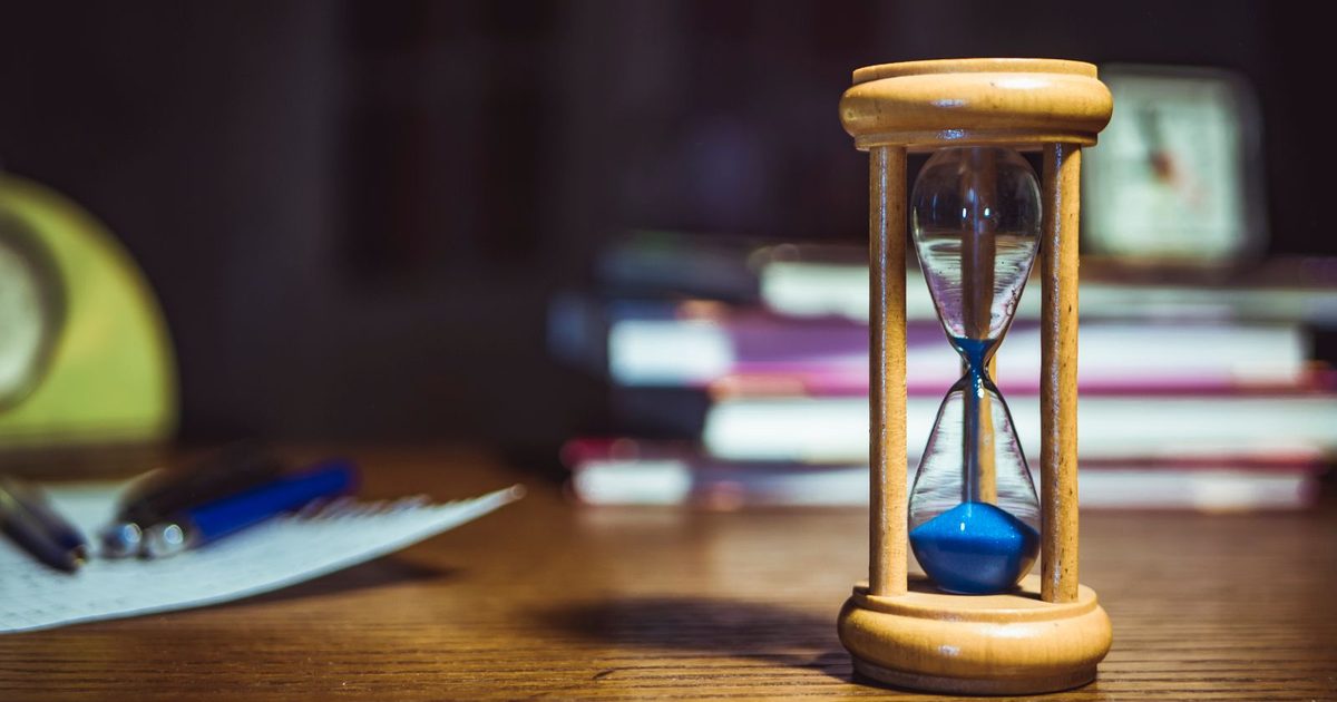 A wooden hourglass with blue sand running through it on a desk, with pens, papers and a stack of books blurred behind it