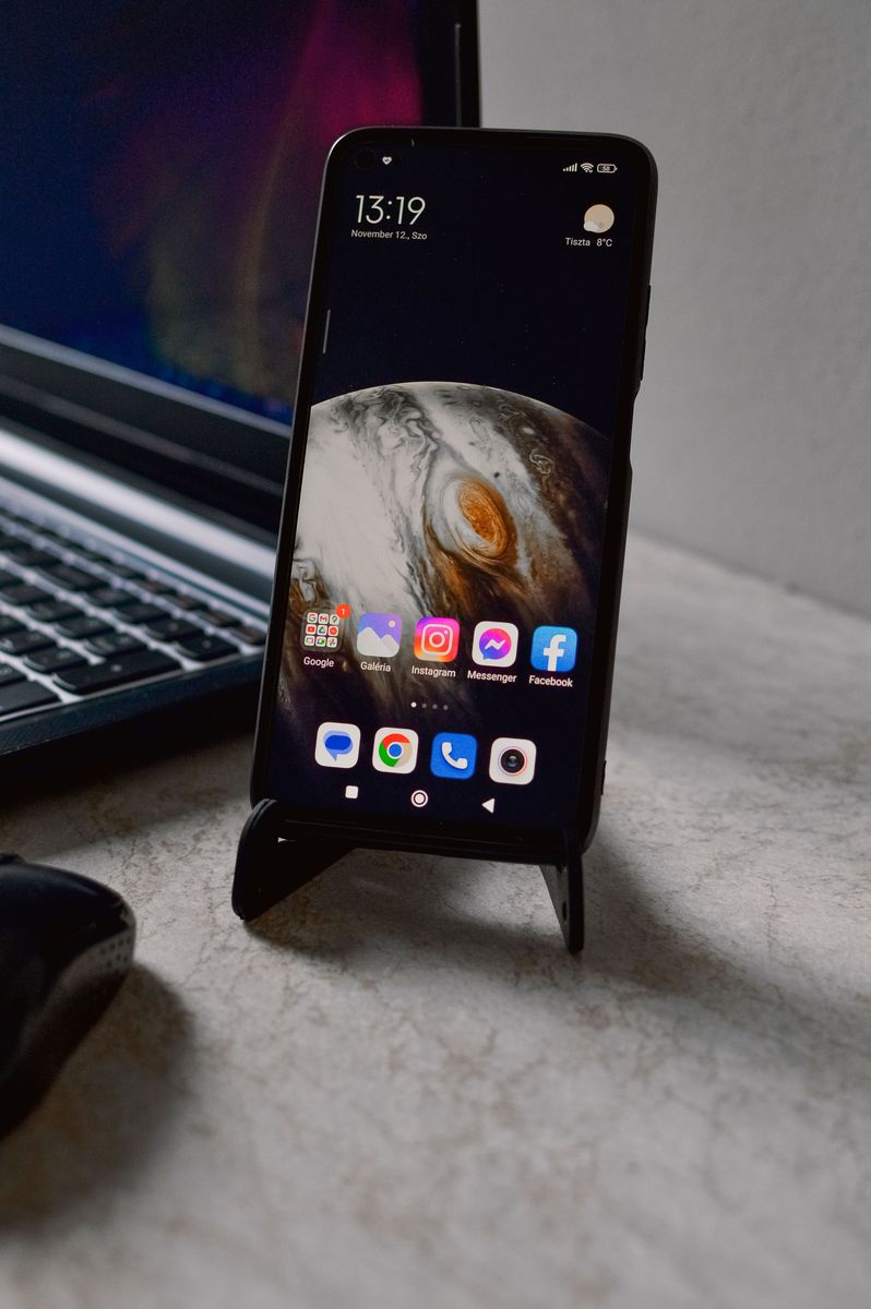 An Android phone on a small stand showing a home screen of app icons, beside an open laptop on a stone desk