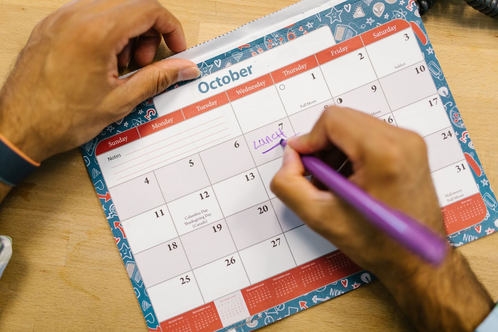 A hand writing a reminder on an October page of a desk calendar with a purple pen