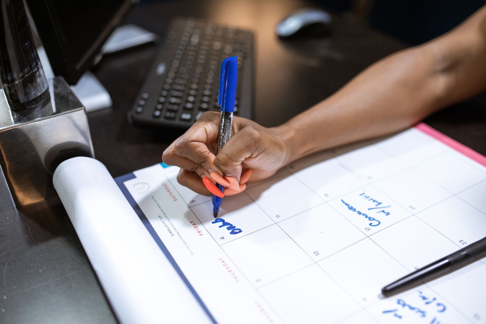 A hand writing a plan in blue marker on a large paper desk calendar beside a keyboard