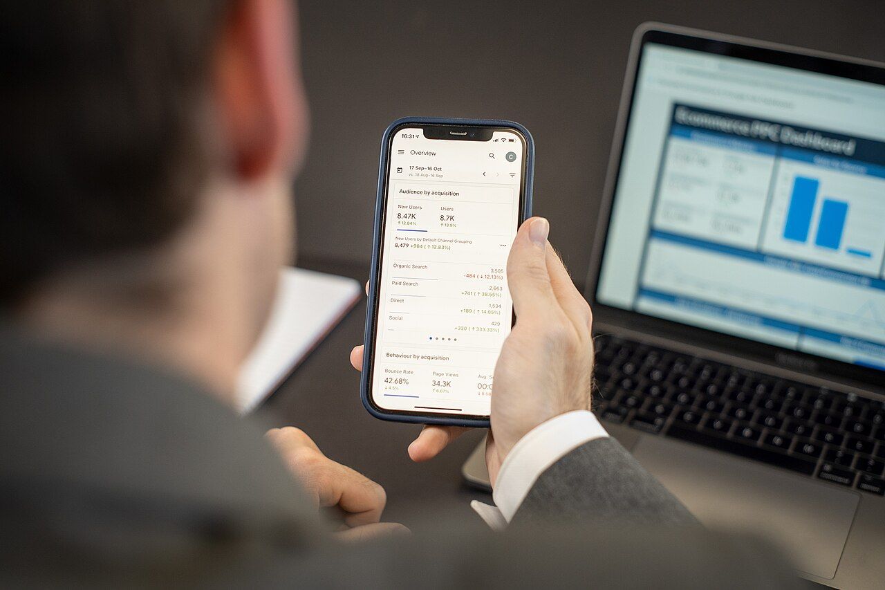 A person in a suit holding a phone showing an analytics dashboard with acquisition numbers, a laptop with a bar chart open behind it