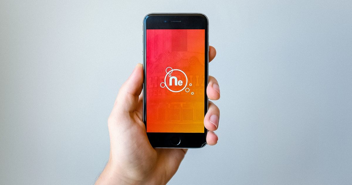 A hand holding an iPhone up against a pale grey wall, its screen showing an orange app splash screen with a round logo