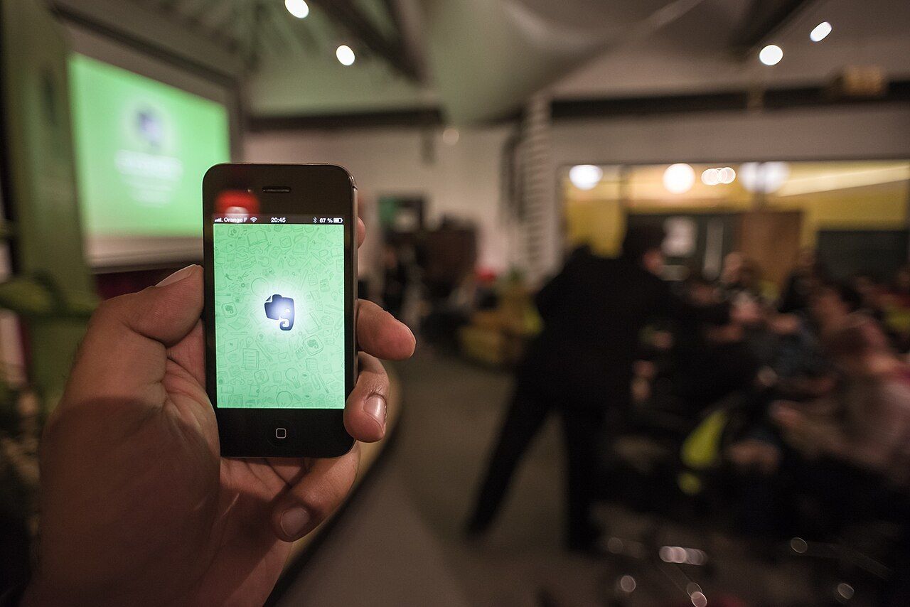 A hand holding up an iPhone showing the Evernote app at a crowded community meetup, with a green slide projected on the wall behind