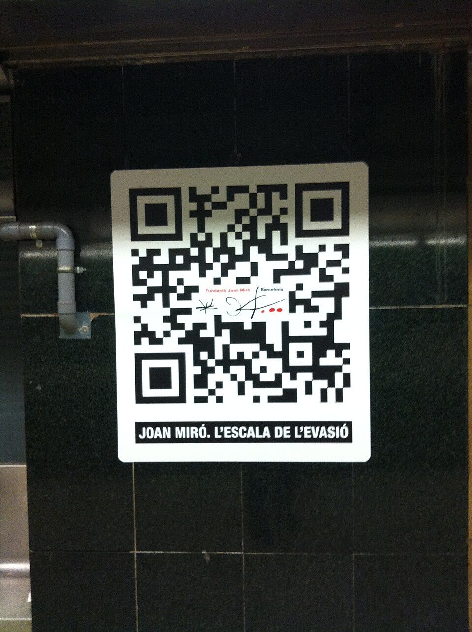 A large black-and-white QR code poster mounted on a dark tiled wall at Plaça de Catalunya in Barcelona, part of a street QR code campaign for a Joan Miró exhibition