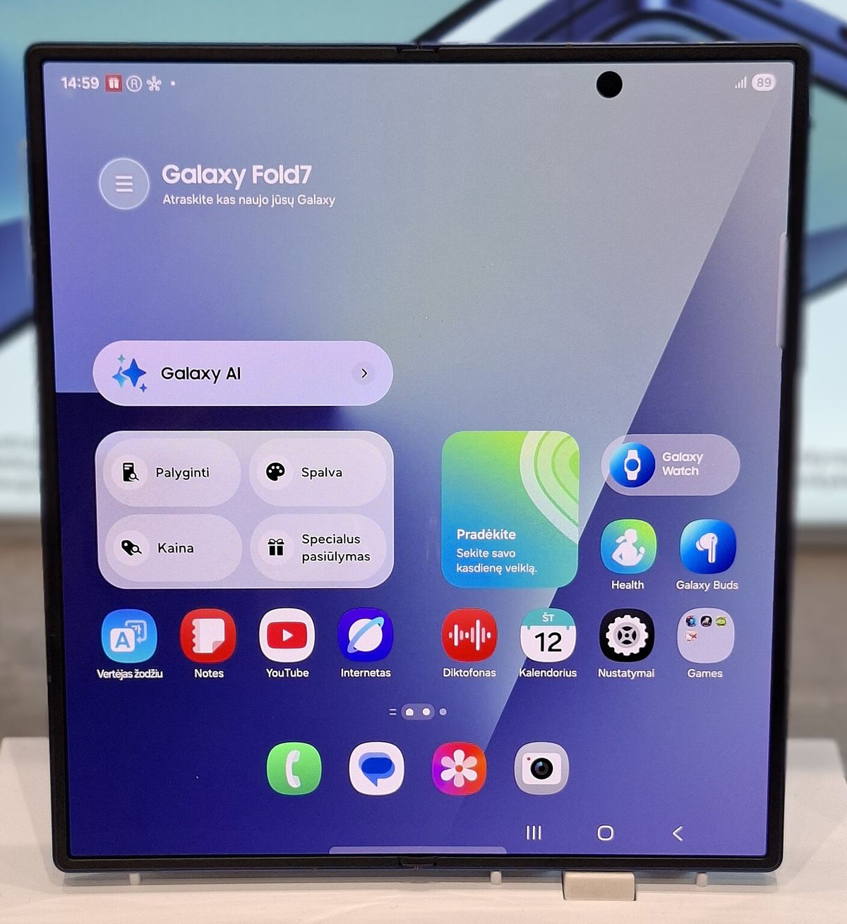 An unfolded book-style foldable phone on a store stand, its wide inner display showing a home screen with widgets and app icons, and a punch-hole front camera near the top right