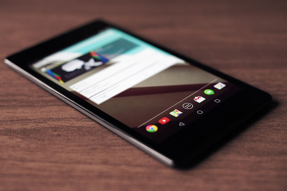 An Android tablet lying on a wooden desk, its home screen showing a weather card and a dock of app icons