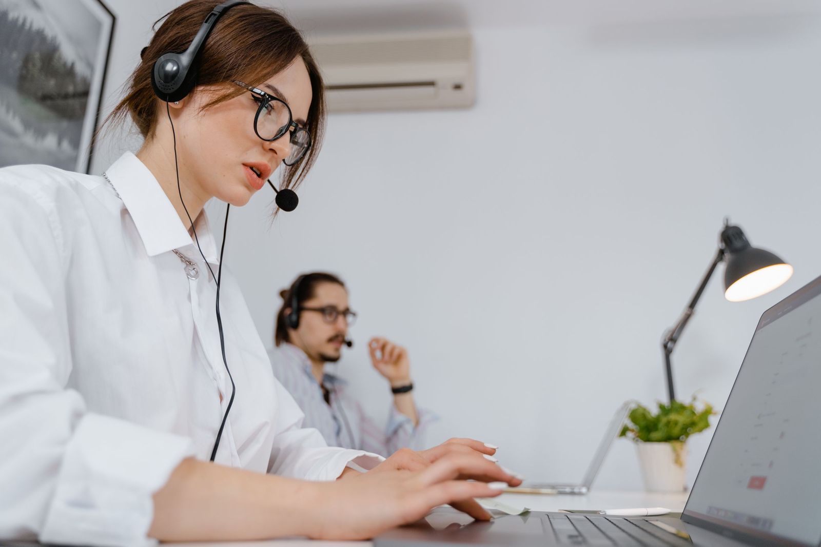A customer support agent wearing a headset typing on a laptop, with a colleague working behind her
