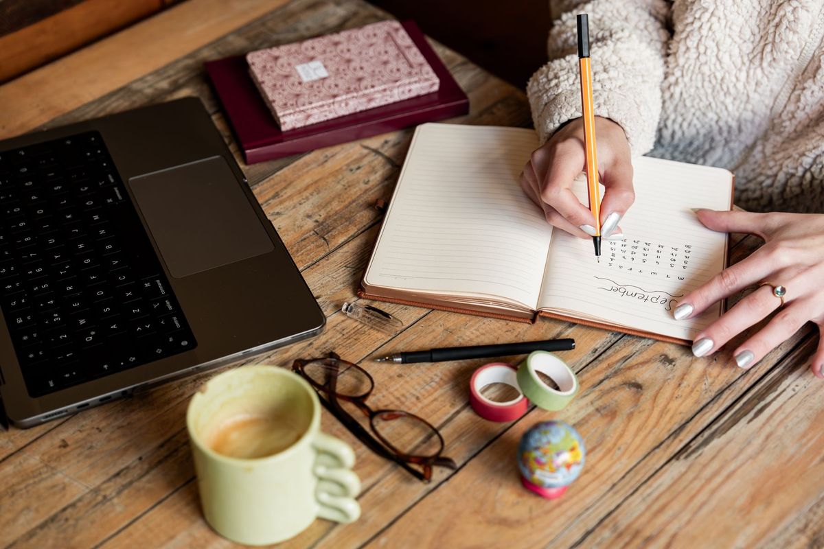 A person writing a plan in a notebook on a wooden desk beside a laptop, a cup of coffee and glasses