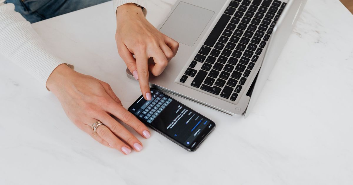 Hands typing a message on a black smartphone lying on a white marble desk next to an open silver laptop