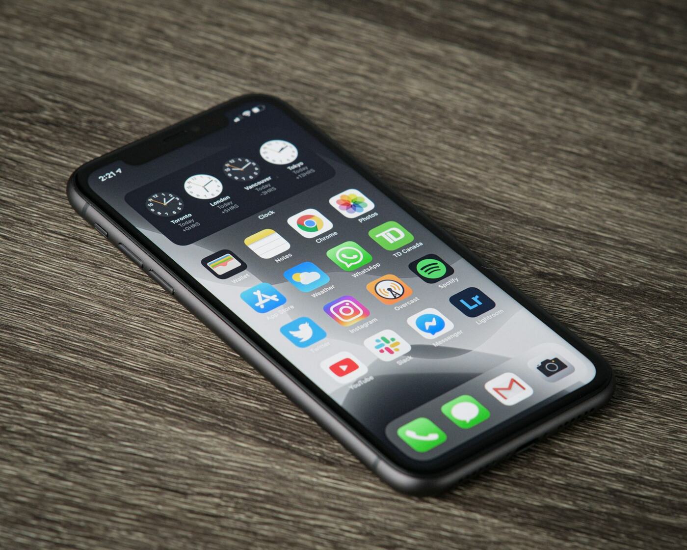 An iPhone lying on a wooden table, its home screen filled with small app icons such as Chrome, WhatsApp, Spotify, Slack and Gmail